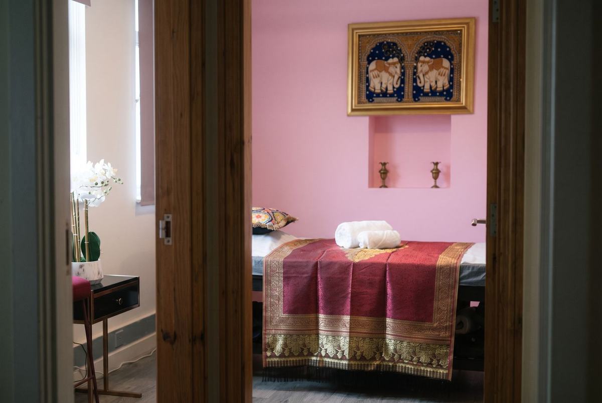 The treatment room seen through an open door, with a silk-dressed massage bed beneath a framed elephant tapestry